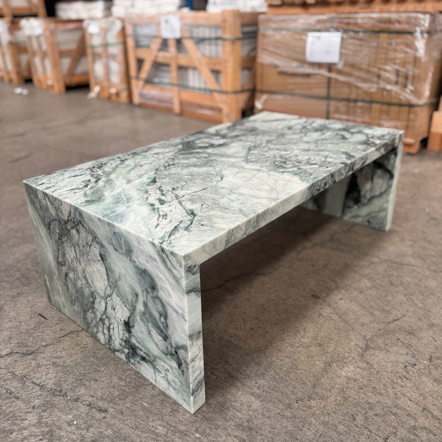 A rectangular coffee table made of green and white marble with dramatic veining, photographed in a warehouse setting with pallets and shelving in the background.